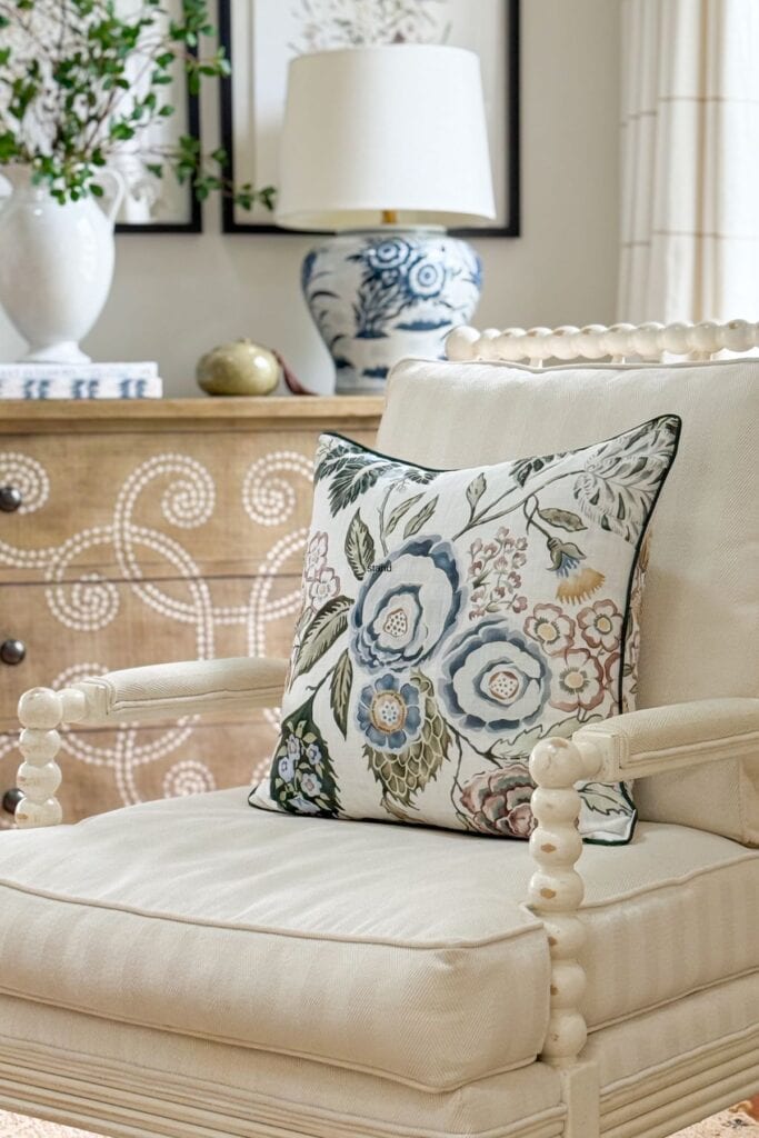 A blue and white ceramic ginger jar table lamp on a natural wood chest in a living room with a cream spool chair and floral pillow in the foreground