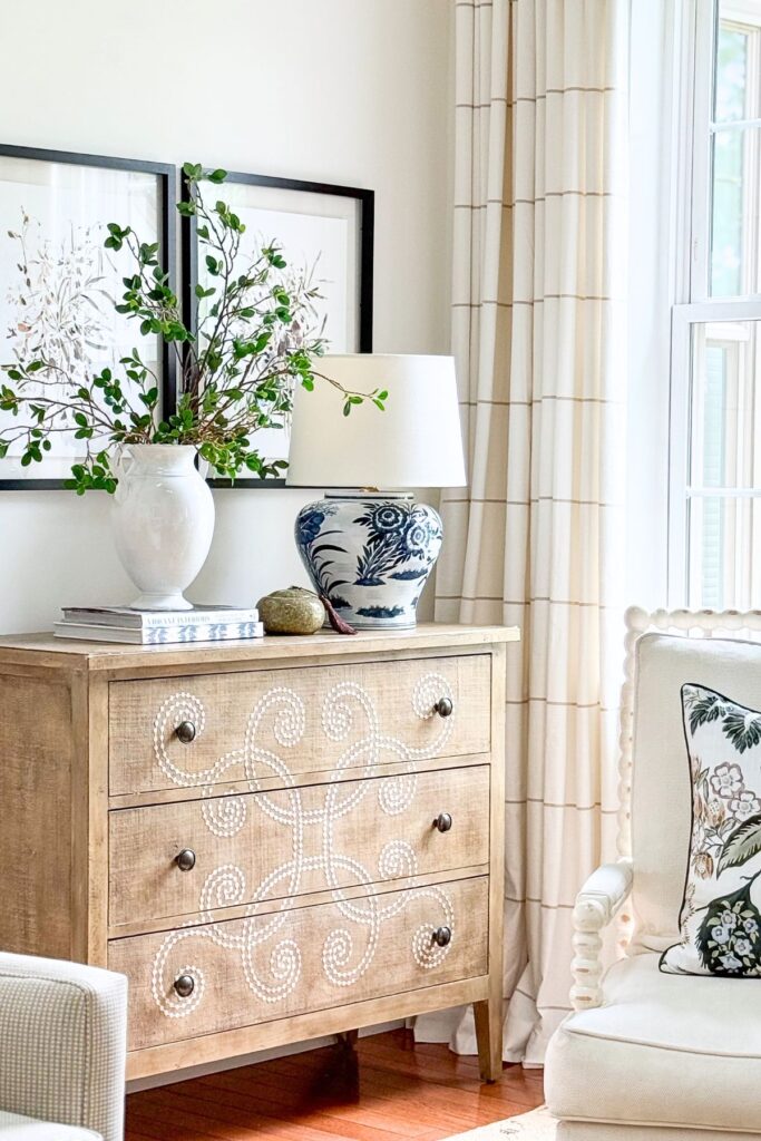 A blue and white ceramic ginger jar table lamp styled on a natural wood chest with a white vase of greenery, stacked books, and framed botanical art