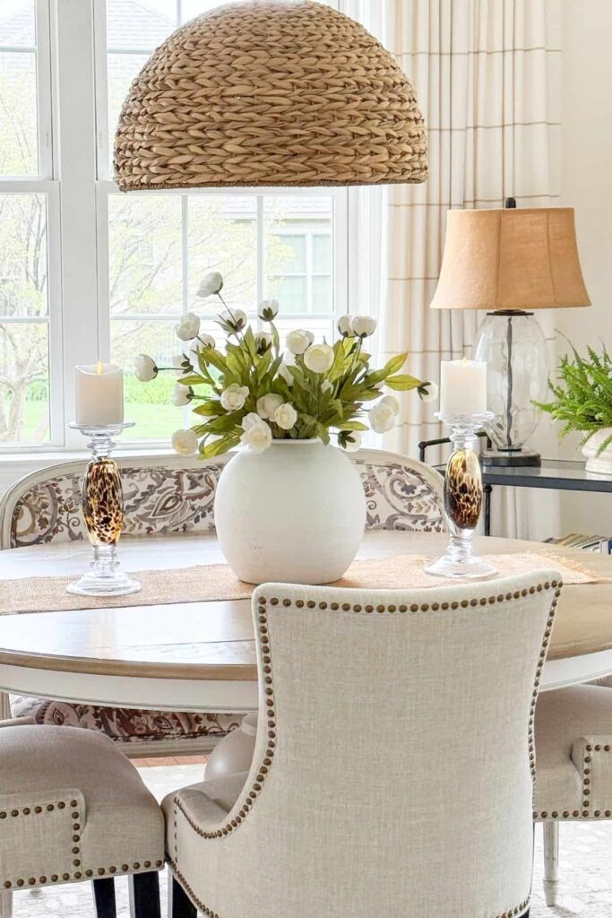 A table lamp with a burlap shade on a black console table in a dining room with a woven pendant light, upholstered chairs, and a round table with white flowers