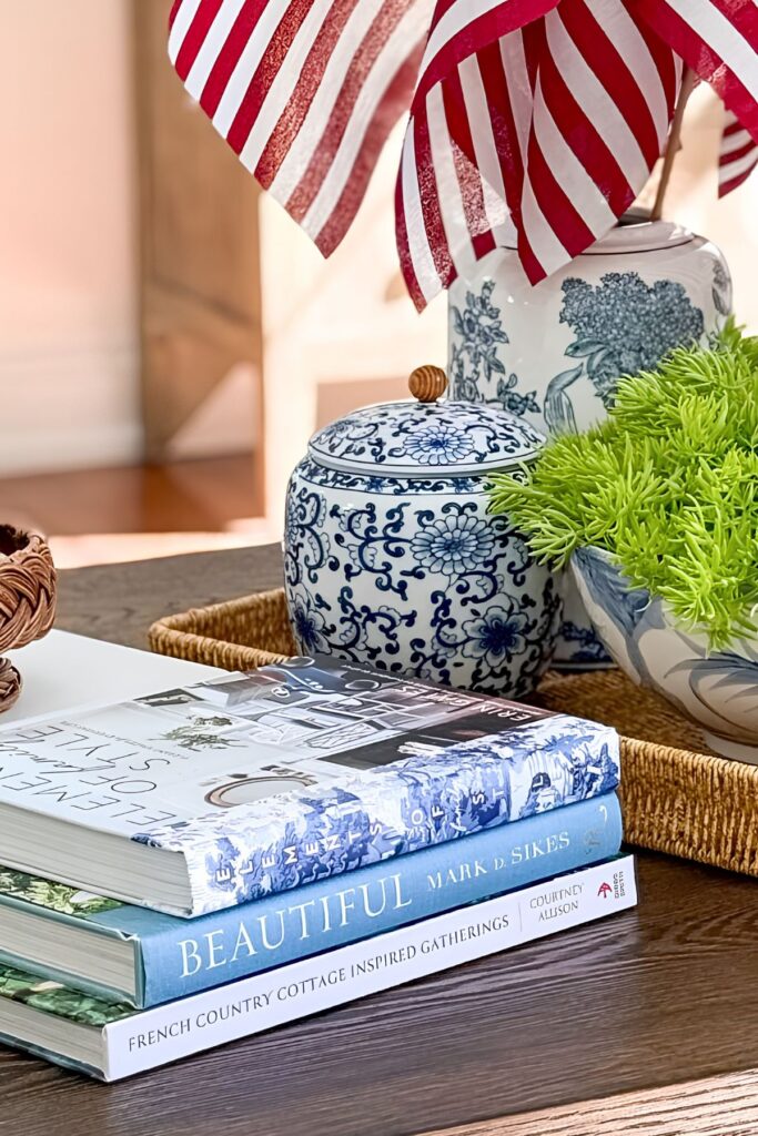 Stacked coffee table books beside a blue and white ginger jar and a bowl of Lemon Coral Sedum with American flags on a wicker tray for a patriotic living room coffee table vignette.