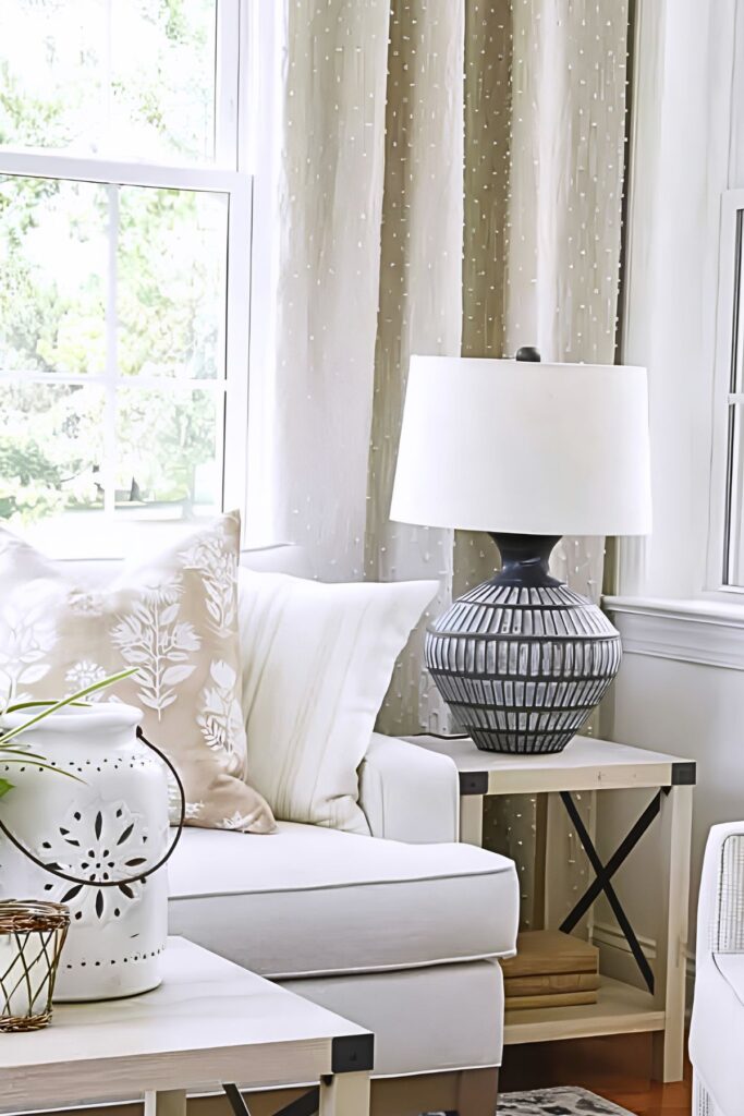 A dark ribbed ceramic table lamp with a white shade on a natural wood end table next to a neutral sofa with decorative pillows in a bright living room