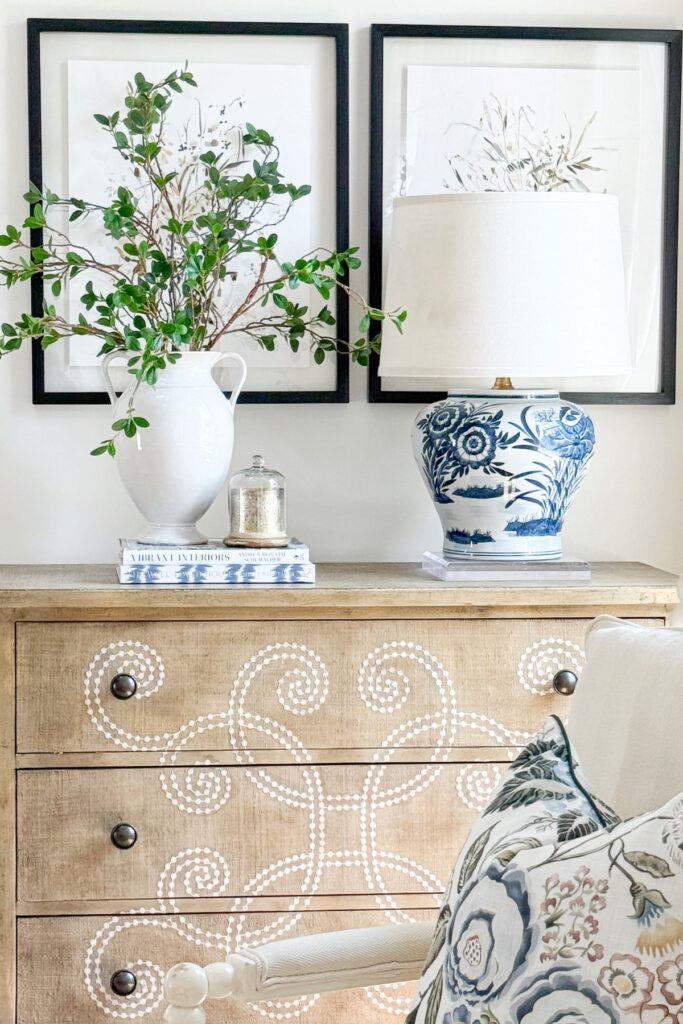 A DIY blue and white chinoiserie ginger jar lamp sitting on a clear acrylic riser on a wood console table styled with a white pitcher, greenery, stacked books, and framed botanical art in a living room.