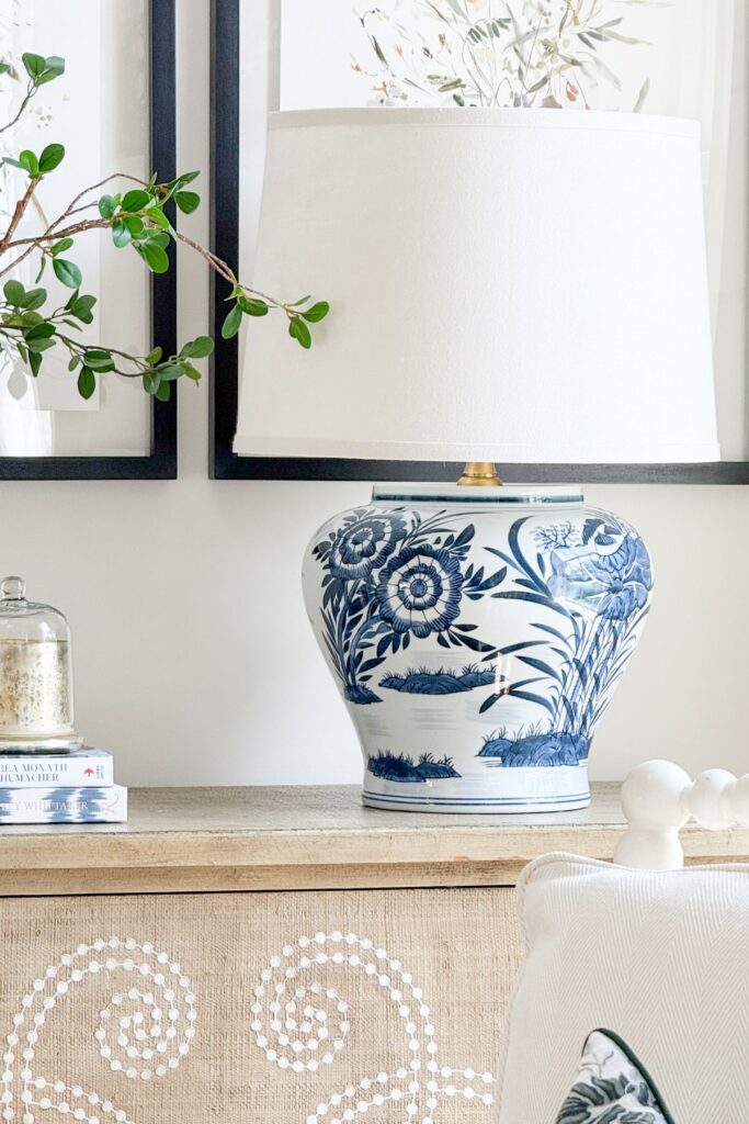 A finished DIY blue and white chinoiserie ginger jar lamp with a white linen shade styled on a wood console table with stacked books and decorative objects in a living room.