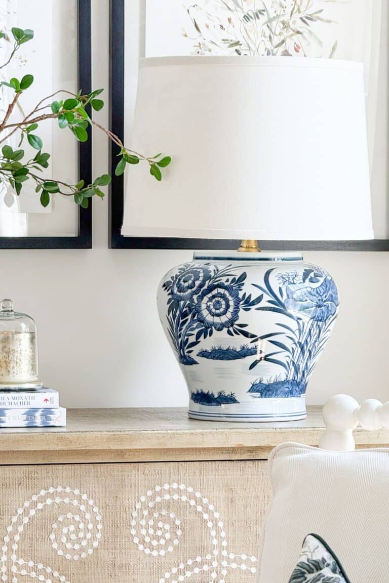 A finished DIY blue and white chinoiserie ginger jar lamp with a white linen shade styled on a wood console table with stacked books and decorative objects in a living room.
