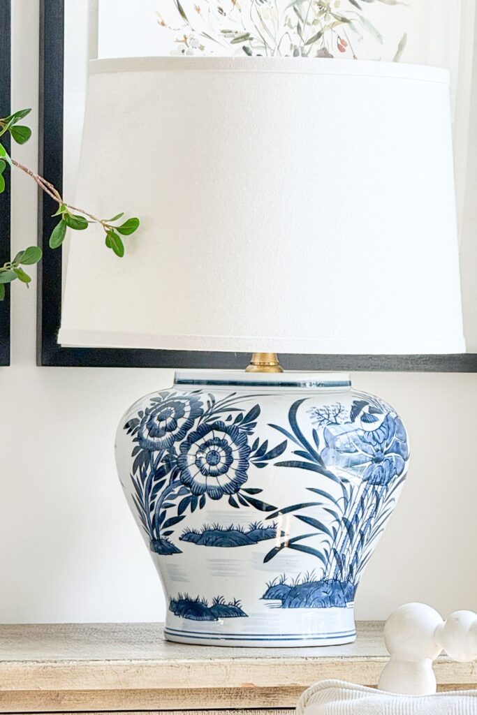 A close up of a finished DIY lamp with a large blue and white chinoiserie ginger jar base and a white linen drum shade on a wood console table.