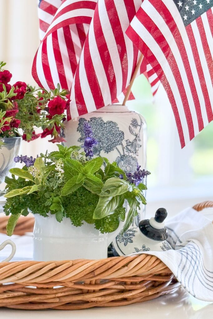 Fresh garden herbs in a small white pitcher beside a tall blue and white chinoiserie ginger jar with American flags and a blue striped tea towel draped over a wicker tray as part of a patriotic kitchen island vignette.