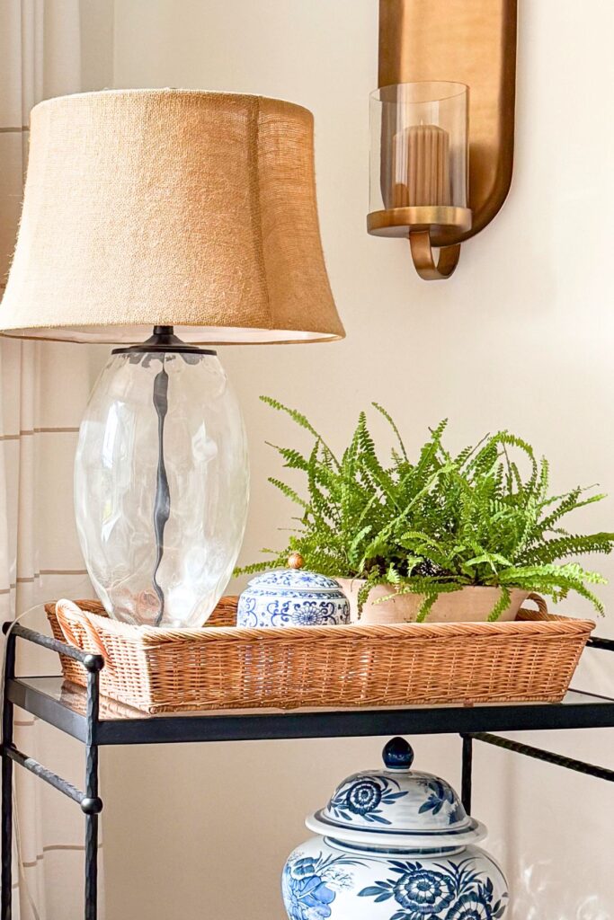 A clear glass table lamp with a burlap shade styled in a wicker tray with a fern and blue and white ginger jar on a black metal bar cart