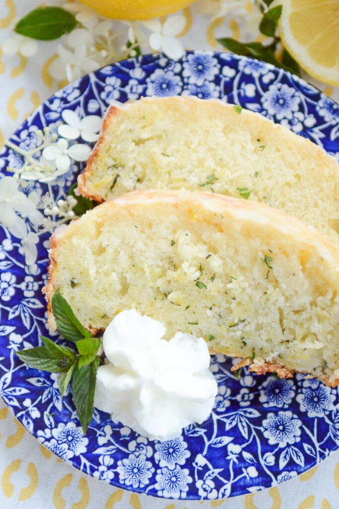 two slices of glazed lemon zucchini bread on a blue and white plate with whipped cream and fresh mint