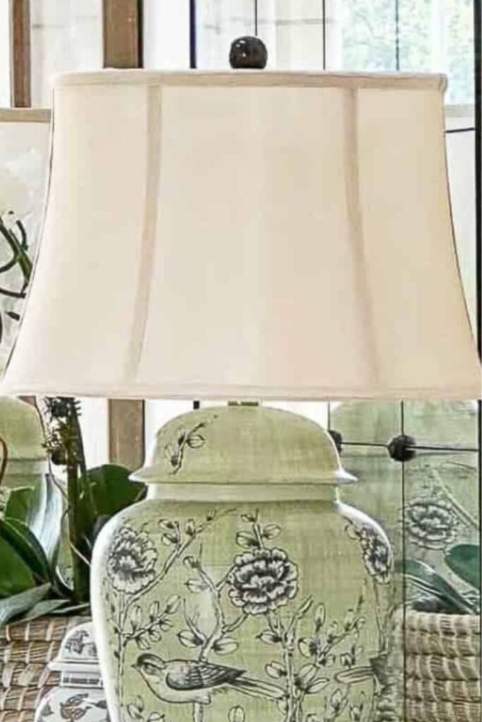 A close up of a green ceramic ginger jar table lamp with hand painted birds and florals and a soft white shade with graceful curved edges