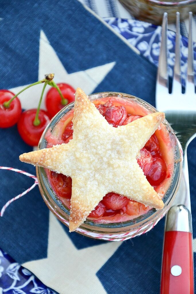 overhead view of a mini cherry pie in a mason jar with a golden star shaped crust topping on a patriotic blue star napkin