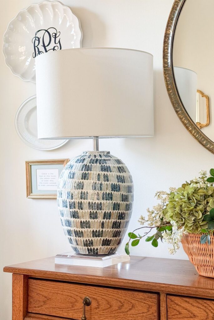 A large mosaic ceramic table lamp on a clear acrylic riser on a wood dresser styled with a wicker basket of hydrangeas and greenery, a monogram plate, and a round gold mirror