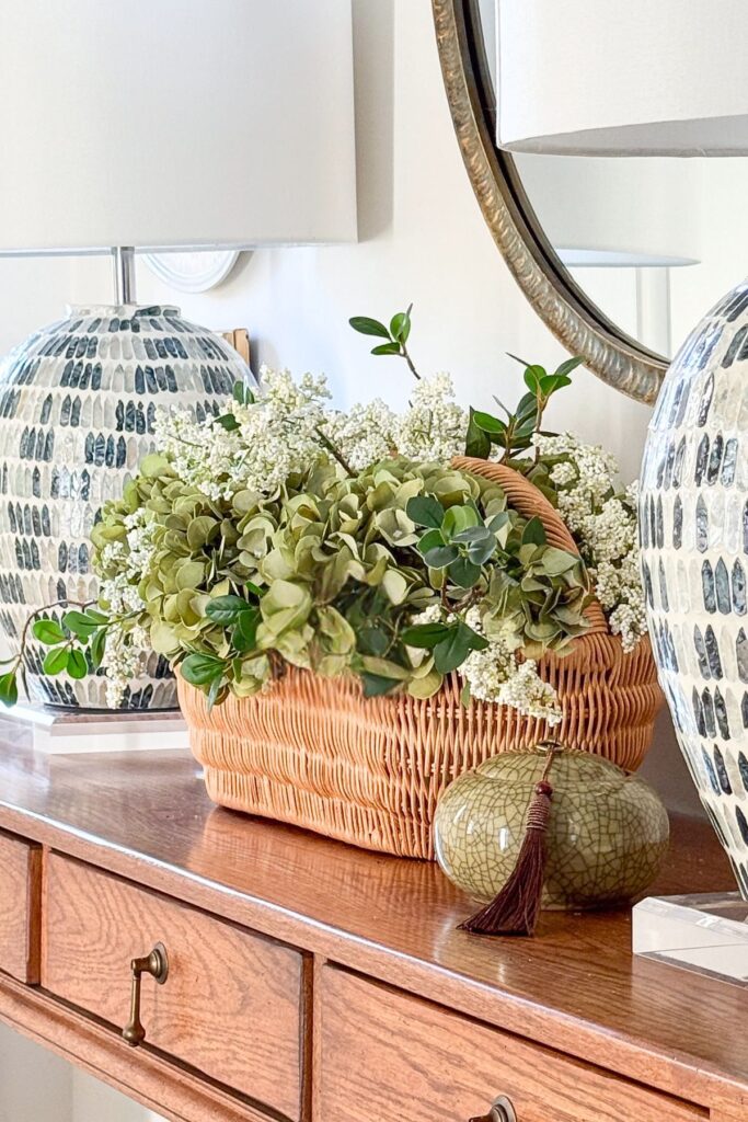 A pair of mosaic ceramic table lamps on a wood console table styled with a wicker basket of hydrangeas and greenery and a round gold mirror
