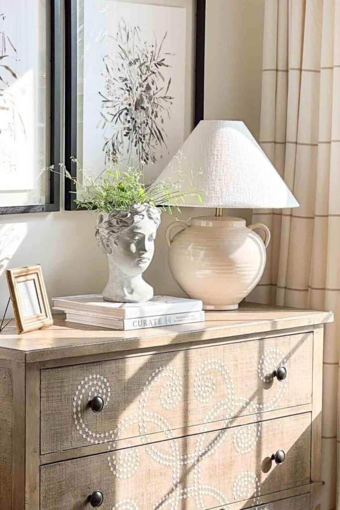 A neutral ceramic urn table lamp with a linen shade styled on a wooden dresser with stacked books, a sculptural head planter, and framed art