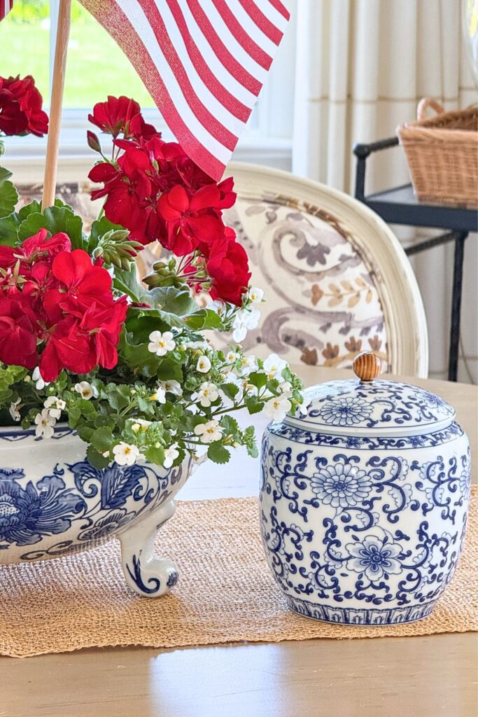 Patriotic dining table arrangement with red geraniums and white Bacopa in a blue and white footed bowl paired with a blue and white ginger jar and American flags.
