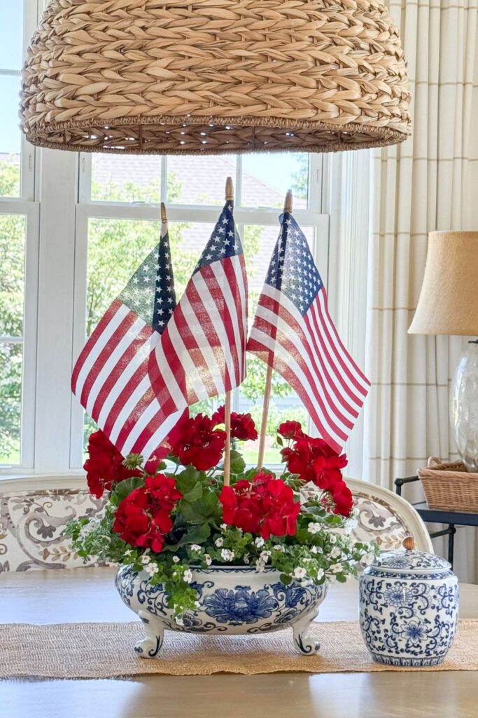 Red geranium and white Bacopa patriotic arrangement with American flags in a blue and white footed bowl on a dining room table beneath a wicker pendant light.