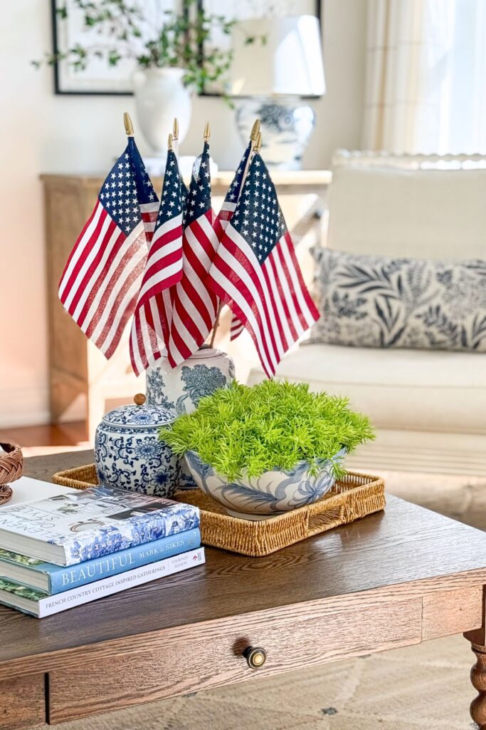 Full living room view of a patriotic coffee table vignette with American flags in a tall blue and white chinoiserie ginger jar, Lemon Coral Sedum in a blue and white bowl, a small ginger jar, and stacked coffee table books on a wicker tray.