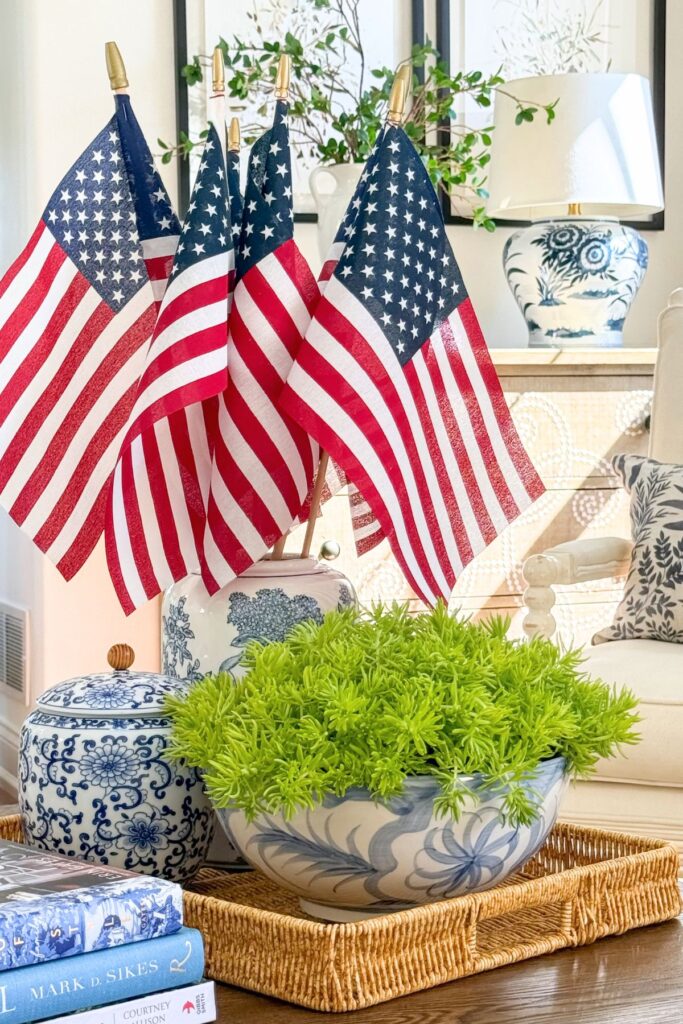 Full living room view of a patriotic coffee table vignette with American flags in a tall blue and white chinoiserie ginger jar, Lemon Coral Sedum in a blue and white bowl, a small ginger jar, and stacked coffee table books on a wicker tray.