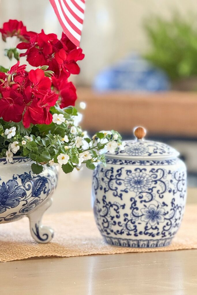 Calliope Large Dark Red geraniums and white Bacopa planted in a blue and white footed bowl with American flags beside a small blue and white ginger jar on a burlap runner for a patriotic dining table arrangement.