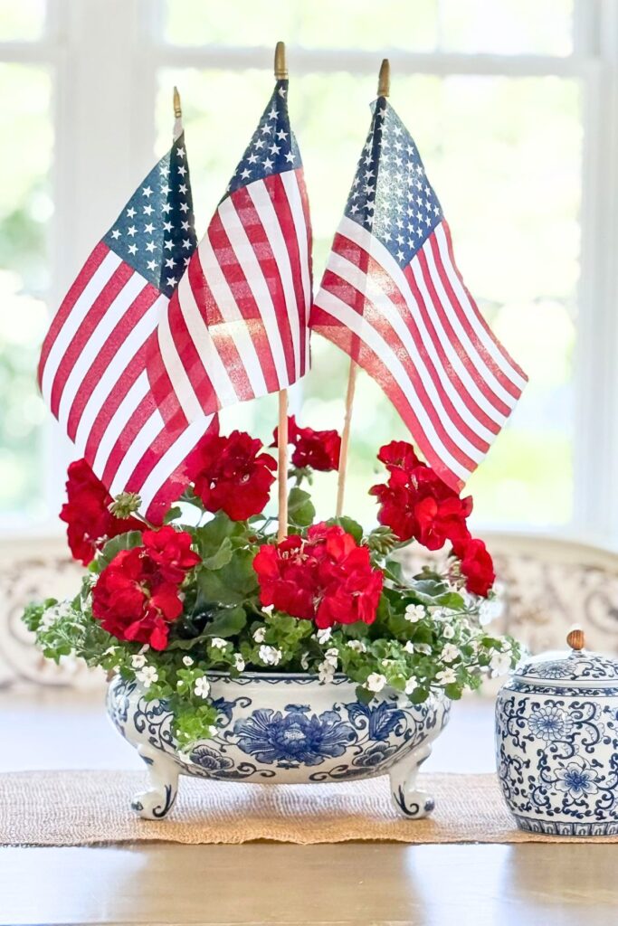 Red geranium and white Bacopa patriotic arrangement with American flags in a blue and white footed bowl on a dining room table beneath a wicker pendant light.