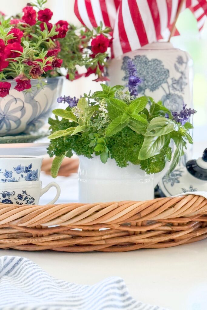 Patriotic kitchen island vignette detail showing stacked vintage blue and white teacups, fresh garden herbs in a white pitcher, a small blue and white teapot, and a blue striped tea towel on a wicker tray with red calibrachoa and American flags in the background.