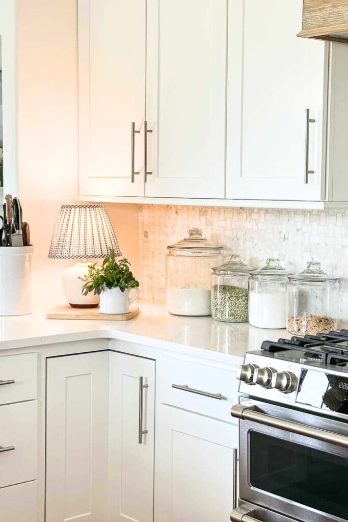 A small ceramic table lamp with a gingham shade glowing warmly on a kitchen counter corner with white cabinets, a marble backsplash, and glass canisters