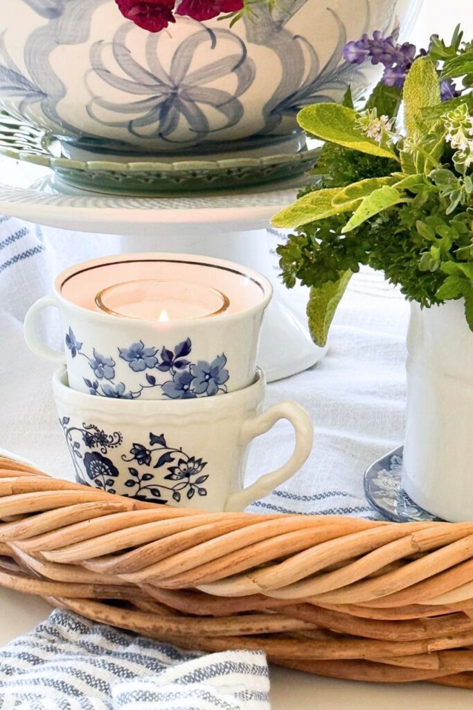 Two stacked mismatched vintage blue and white teacups with a glowing votive candle in the top cup on a wicker tray beside a white cake stand holding a blue and white bowl of red calibrachoa and fresh garden herbs in a white pitcher.