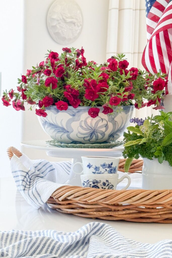 Superbells Double Ruby calibrachoa planted in a blue and white bowl elevated on a white cake stand as part of a patriotic kitchen island vignette with American flags and fresh garden herbs.