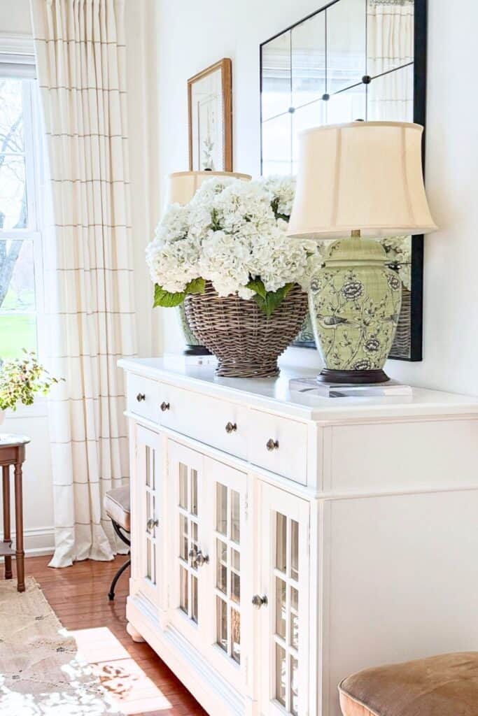 Faux white hydrangeas in a woven basket styled on a white buffet beside a green chinoiserie lamp for a midsummer decorating refresh