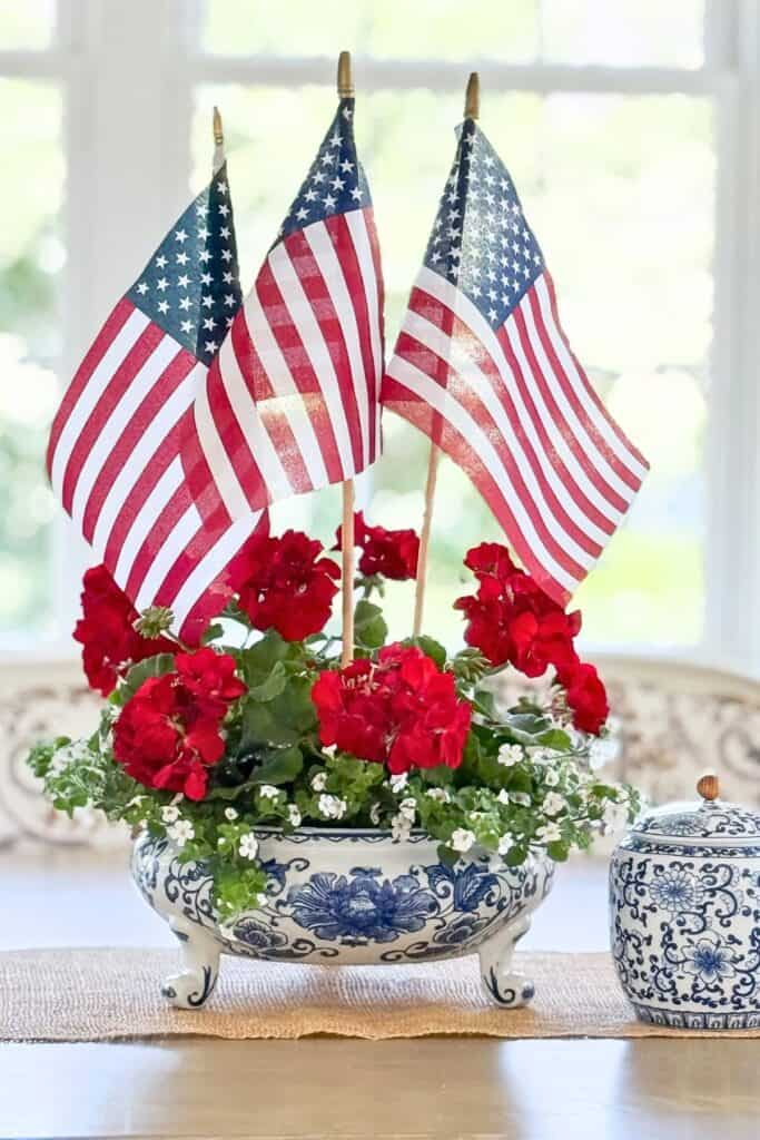 Red geraniums and American flags arranged in a blue and white chinoiserie planter on a dining room table before a midsummer decor refresh