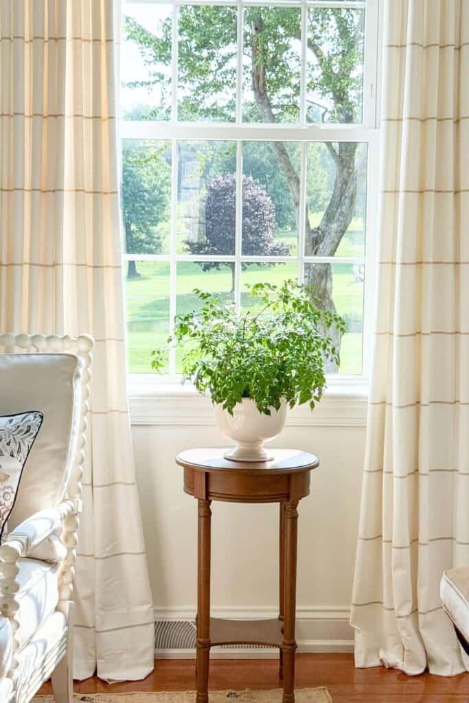 Lush green plant in a white footed urn styled on a side table in front of a window for a midsummer decorating refresh