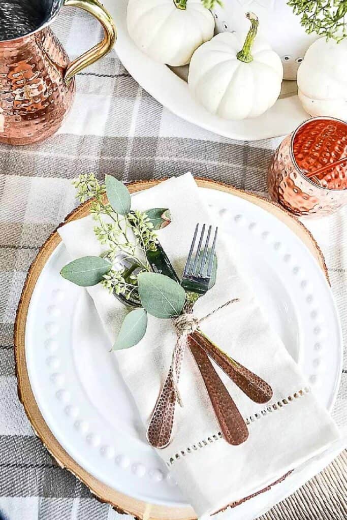 Fall table place setting with a wood slab charger, white plate, linen napkin tied with eucalyptus and copper flatware, and white pumpkins