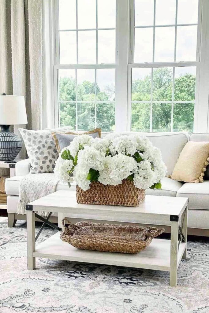 Basket of white faux hydrangeas on a coffee table in front of a light gray sofa with patterned pillows