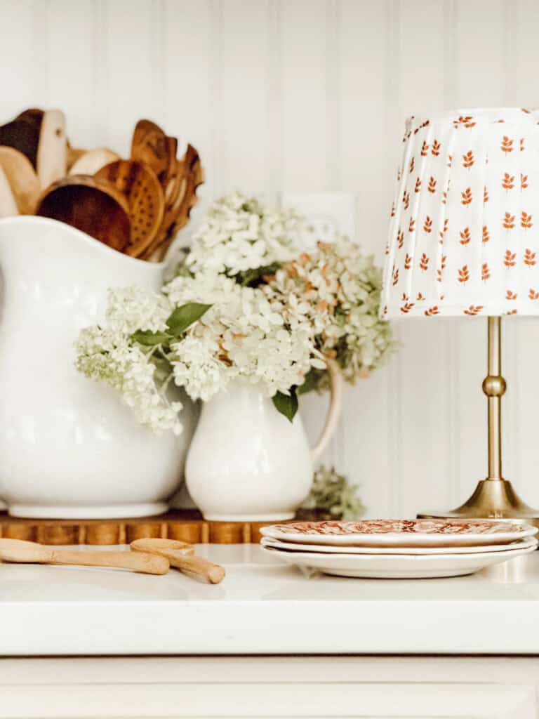 CLOSE UP OF A LAMP WITH A FABRIC LAMPSHADE AND A CROCK OF WOODEN UTINSELS.