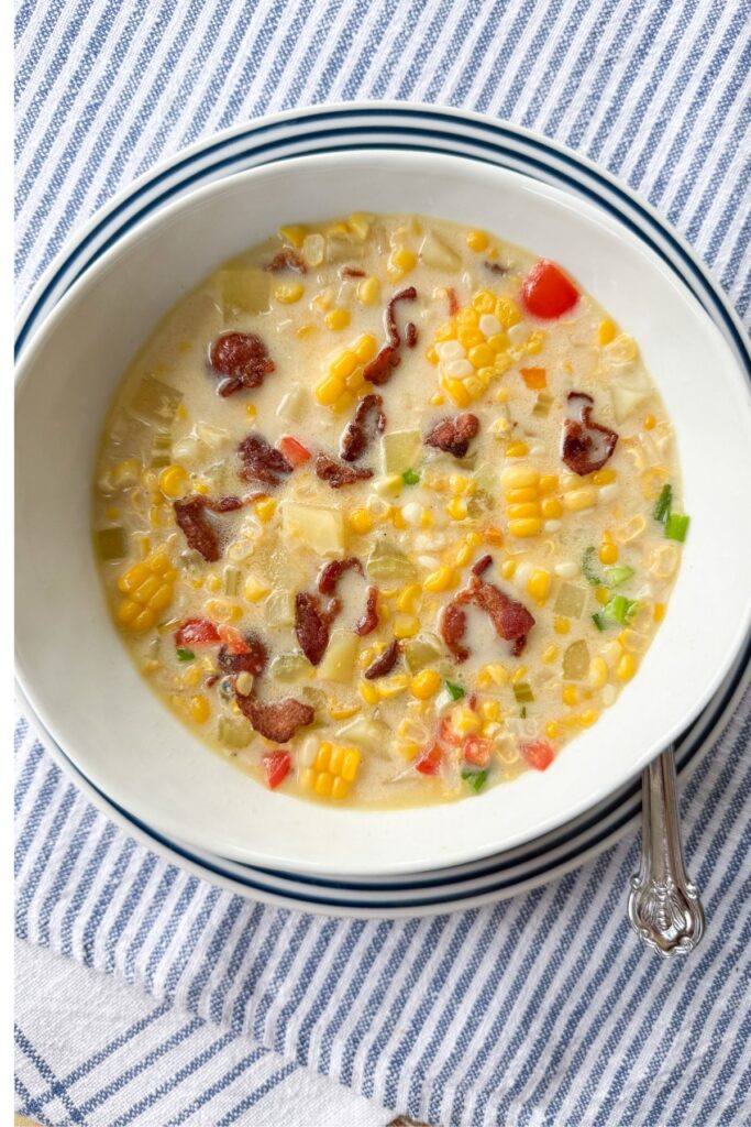 Overhead view of corn chowder with bacon, potatoes, and bell pepper in a white bowl on blue striped linen with a vintage spoon