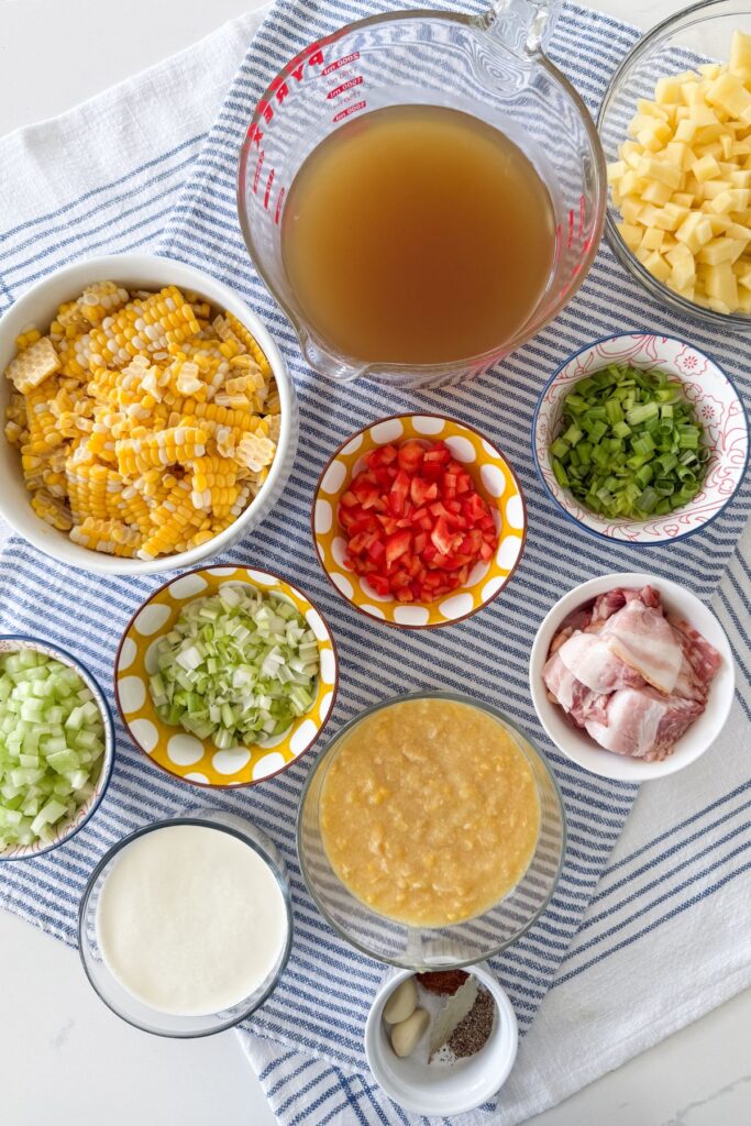 Corn chowder ingredients including fresh corn, chicken stock, potatoes, bell pepper, celery, green onion, creamed corn, bacon, and cream in bowls