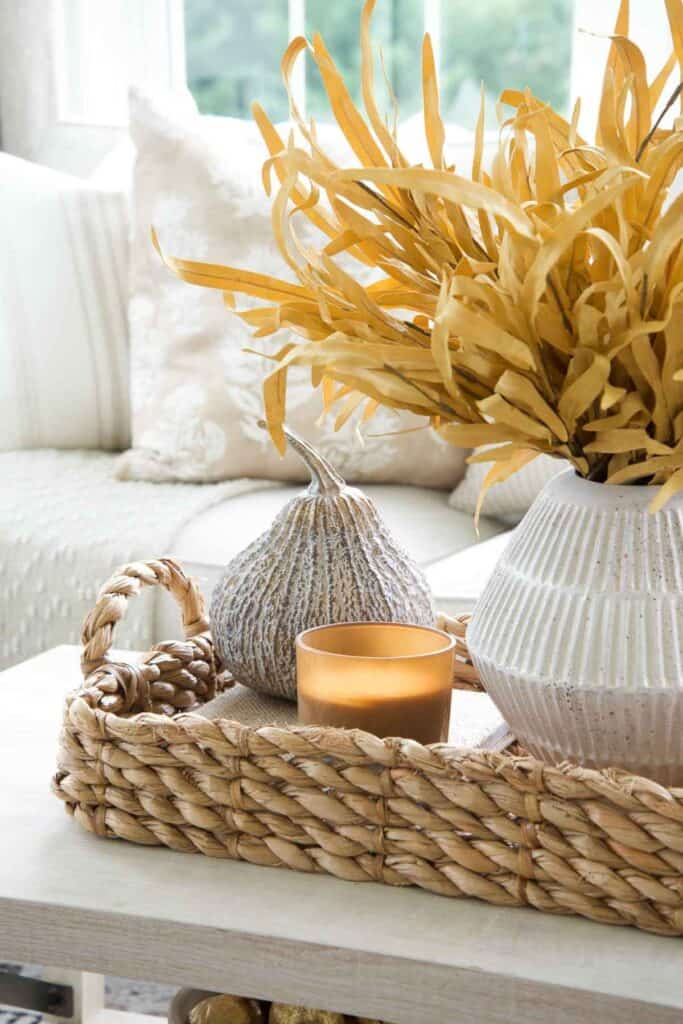 Fall centerpiece on a coffee table with dried corn stalks in a white vase, a stone pumpkin, and an amber candle in a woven tray