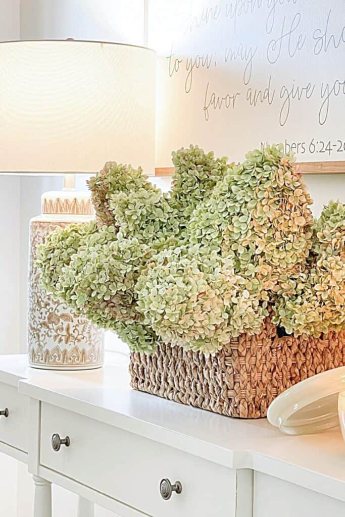 Woven basket of dried green and blush hydrangeas on a white foyer table beside a patterned lamp