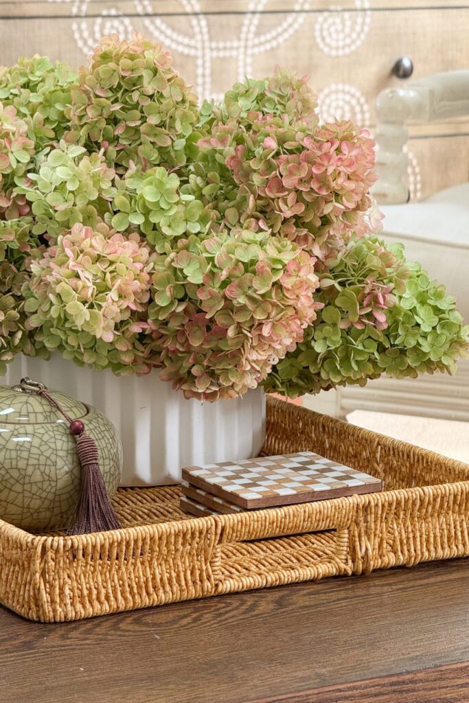 Dried green and pink panicle hydrangeas in a white fluted vase styled on a woven tray with a checkered coaster stack