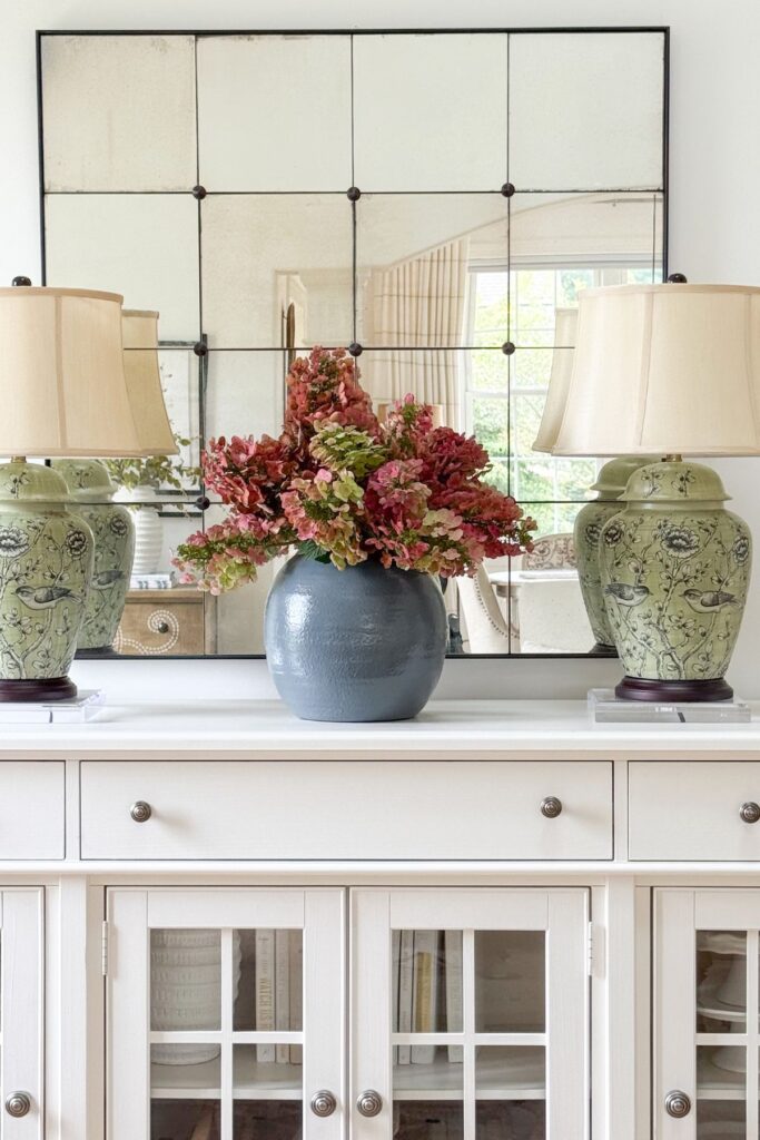 Dried pink and green hydrangeas in a matte blue vase on a white buffet flanked by green chinoiserie lamps