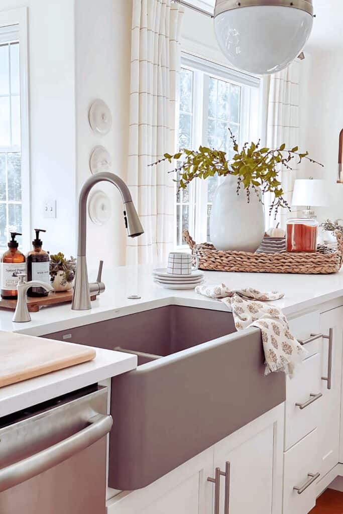 Fall kitchen counter with a gray farmhouse sink, a white vase of greenery, and a woven tray with a candle and mini pumpkins