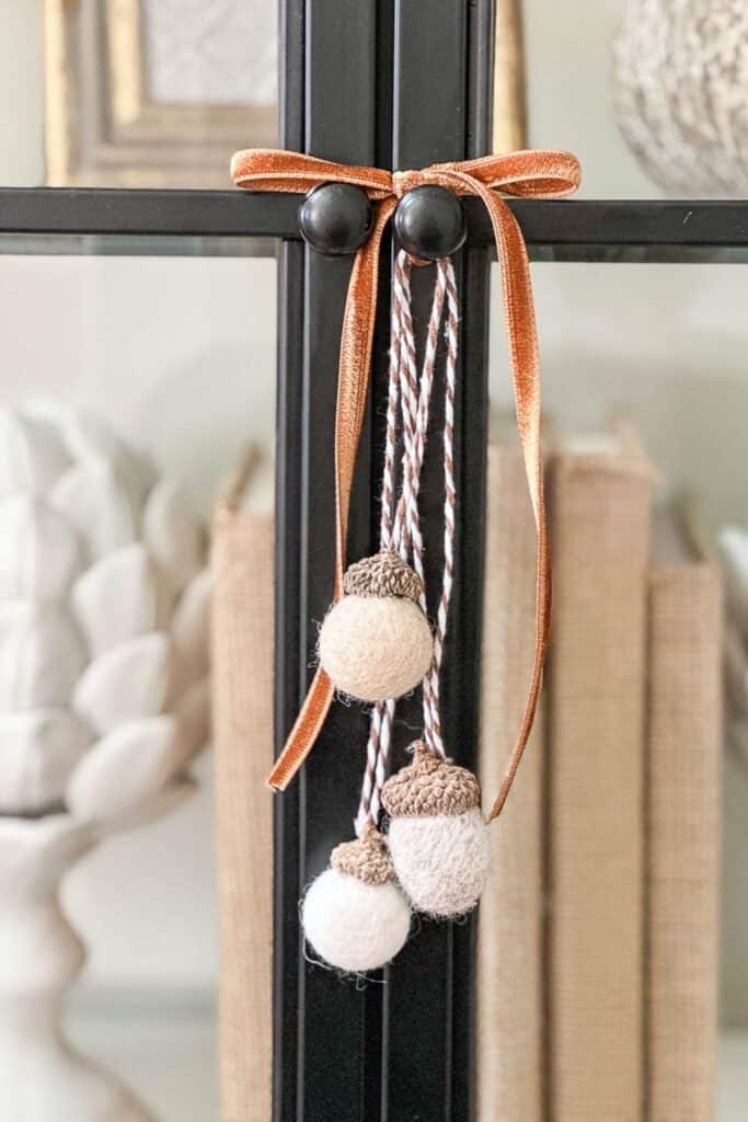 Felted wool ball acorns tied with velvet ribbon and twine hanging from a black bookcase for a fall DIY