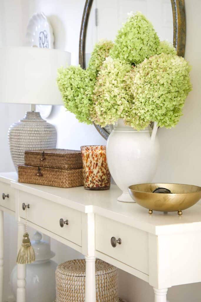 Green and blush hydrangeas in a white pitcher on a white entry console table with woven boxes and a gold bowl