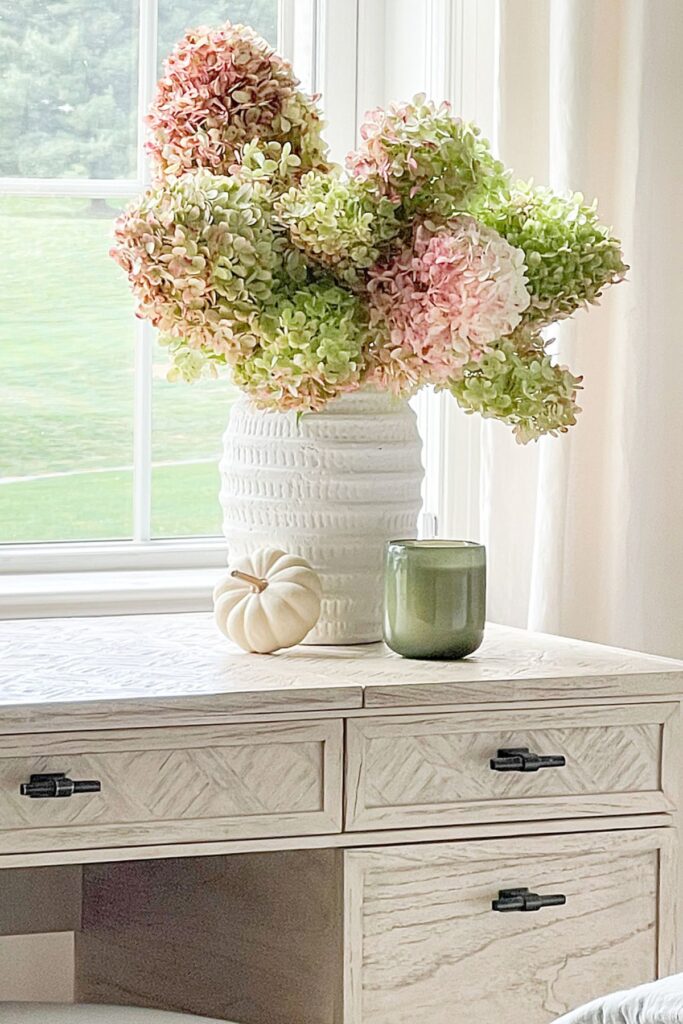 Pink and green drying hydrangeas in a textured white vase on a bedroom dresser beside a window