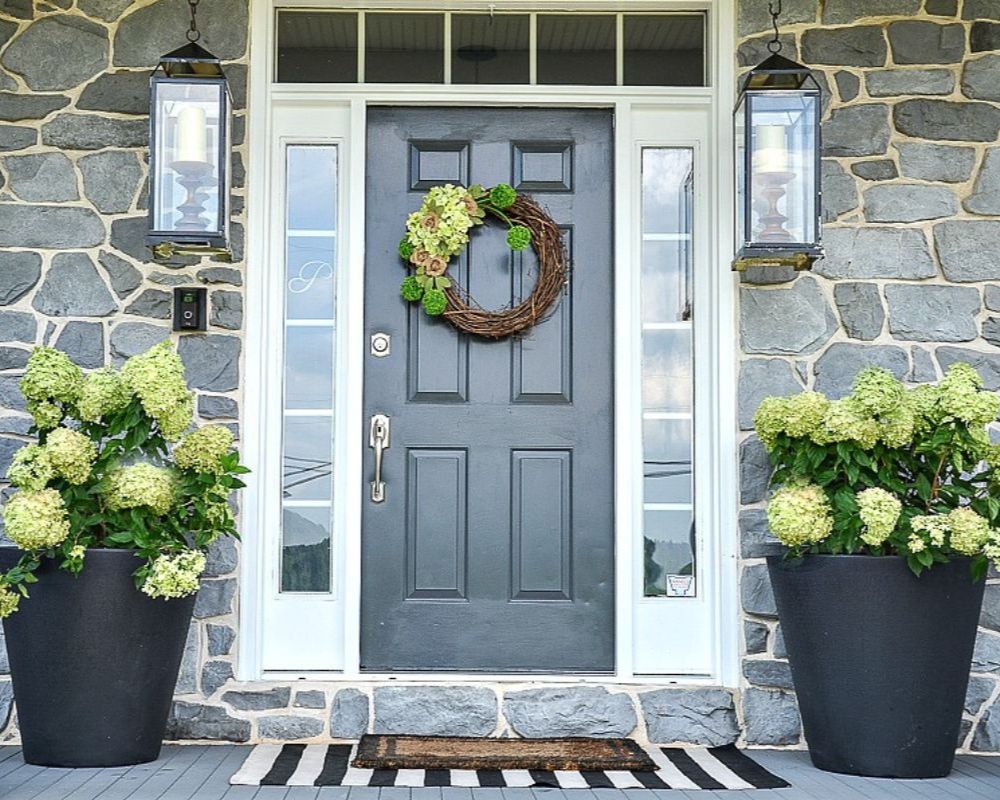 Potted green hydrangeas in black planters flanking a front door with a grapevine hydrangea wreath