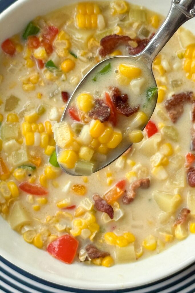 Spoonful of creamy corn chowder with fresh corn, bacon, potatoes, and bell pepper lifted from a white bowl