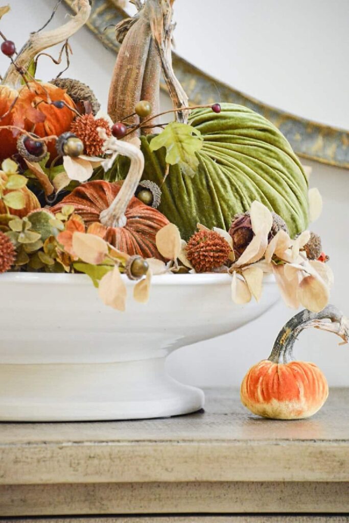 Velvet pumpkins in orange and green with acorns and dried hydrangeas in a white pedestal bowl for a fall bucket list