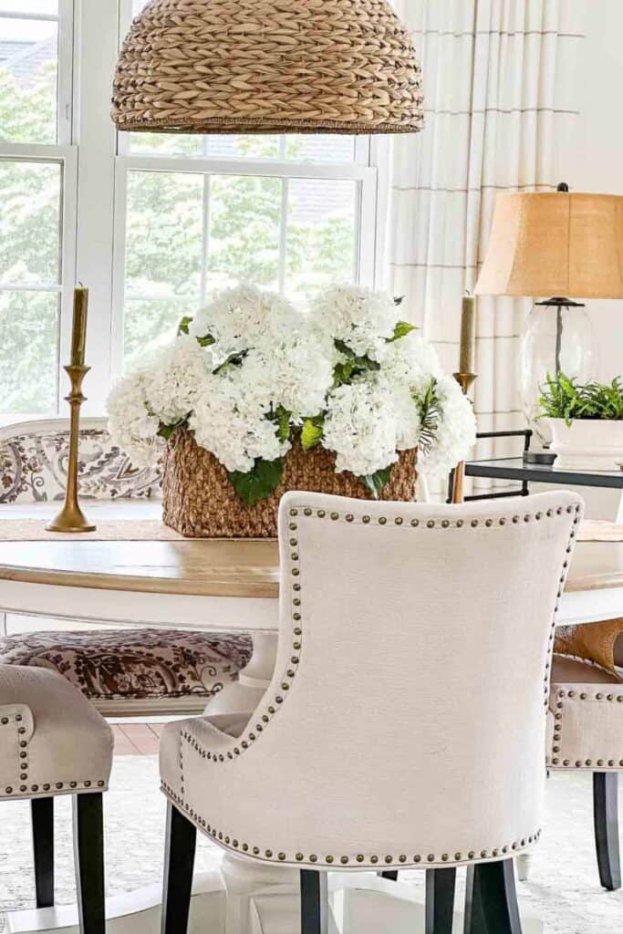 Woven basket filled with white faux hydrangeas as a centerpiece on a dining table with nailhead upholstered chairs
