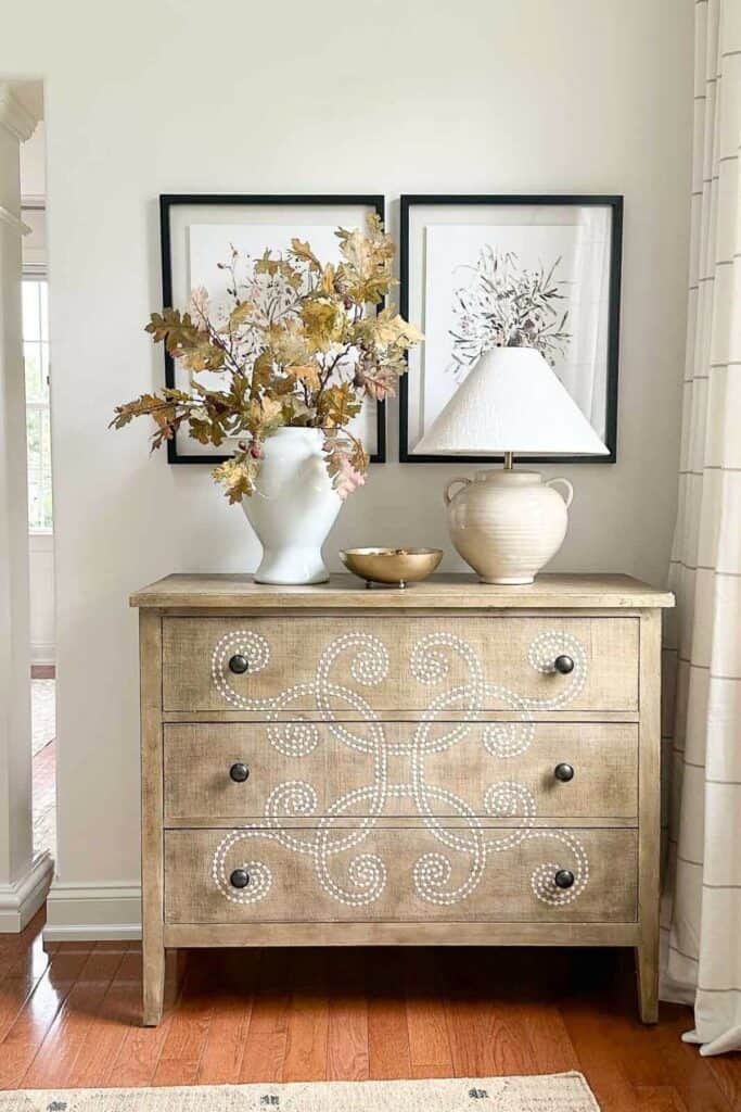 White urn filled with fall oak branches on a curved chest with framed botanical art and a cream lamp, decorated without pumpkins