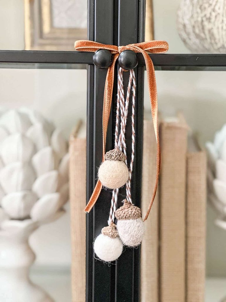 Felted wool ball acorns in cream and oatmeal tones hanging from a copper velvet bow and brown and white twine on a black glass cabinet door