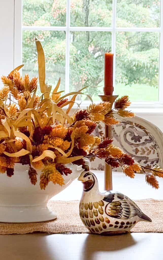 ceramic partridge figurine beside a fall floral centerpiece and taper candle in a window
